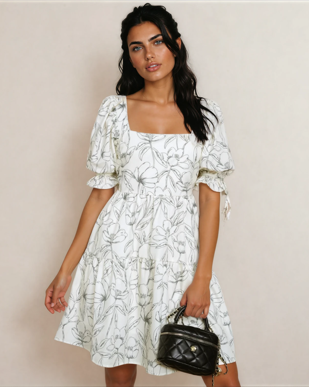 A woman wearing a cream-colored, flowy floral print mini dress with short puff sleeves, a square neckline, and a flattering shirred back detail.
