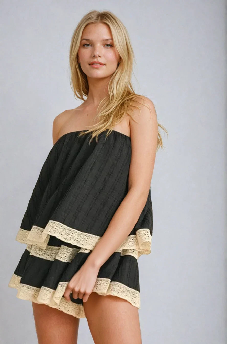 A woman wearing a black tiered strapless tube romper with cream lace trim along the edges, designed with a flowy silhouette and hidden built-in shorts.