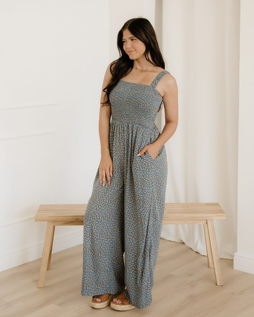 A woman wearing a sleeveless blue floral high-waist jumpsuit featuring an elegant square neckline and a comfortable smocked bodice.