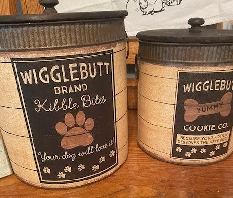 Two rustic metal dog treat canisters side-by-side featuring vintage typography; the larger bin reads Wigglebutt Brand Kibble Bites and the smaller bin reads Wigglebutt Brand Yummy Cookie Co.