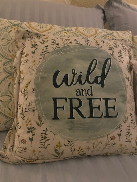 Rustic square accent pillow featuring a vintage-style floral pattern background with the words 'wild and free' printed in a bold, stylized black font, styled on a cozy bed.