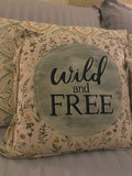 Rustic square accent pillow featuring a vintage-style floral pattern background with the words 'wild and free' printed in a bold, stylized black font, styled on a cozy bed.