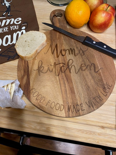 Round wooden cutting board with a handle, intricately engraved with a retro circular design that reads 'Mom's Kitchen — Good Food Made With Love', styled on a kitchen countertop next to fresh fruit