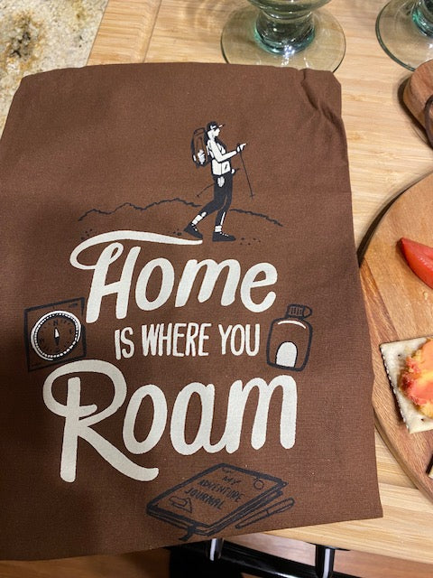 Brown fabric tea towel printed with a retro cream-colored camper graphic and the stylized text 'Home is where you Roam,' displayed flat on a wooden surface.