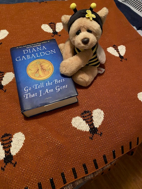 Earthy terracotta woven bee throw blanket styled on a dark wood surface next to a vintage-style teddy bear and book.