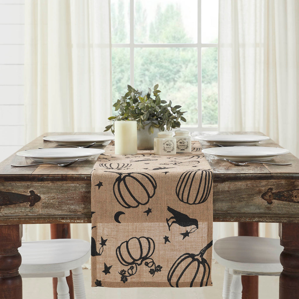 Rustic dining table setup with a beige table runner patterned with black ravens, pumpkins, and crescent moons.