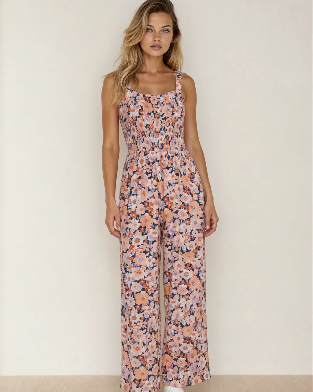 A woman wearing a sleeveless pink floral high-waist jumpsuit with a square neckline and smocked bodice detail.
