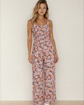A woman wearing a sleeveless pink floral high-waist jumpsuit with a square neckline and smocked bodice detail.

