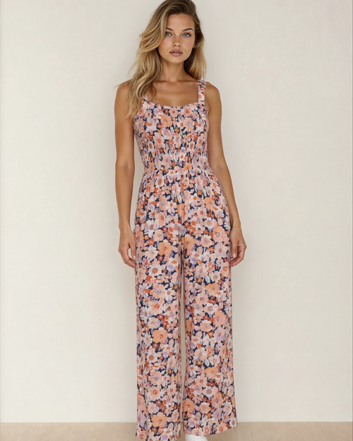 A woman wearing a sleeveless pink floral high-waist jumpsuit with a square neckline and smocked bodice detail.
