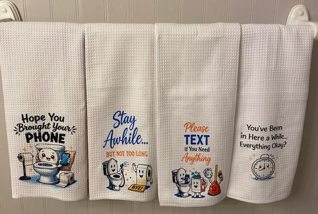 White fabric tea towel featuring a funny illustration of a cartoon cat sitting on a toilet looking at a smartphone, with the text "Hope You Brought Your Phone" written above.