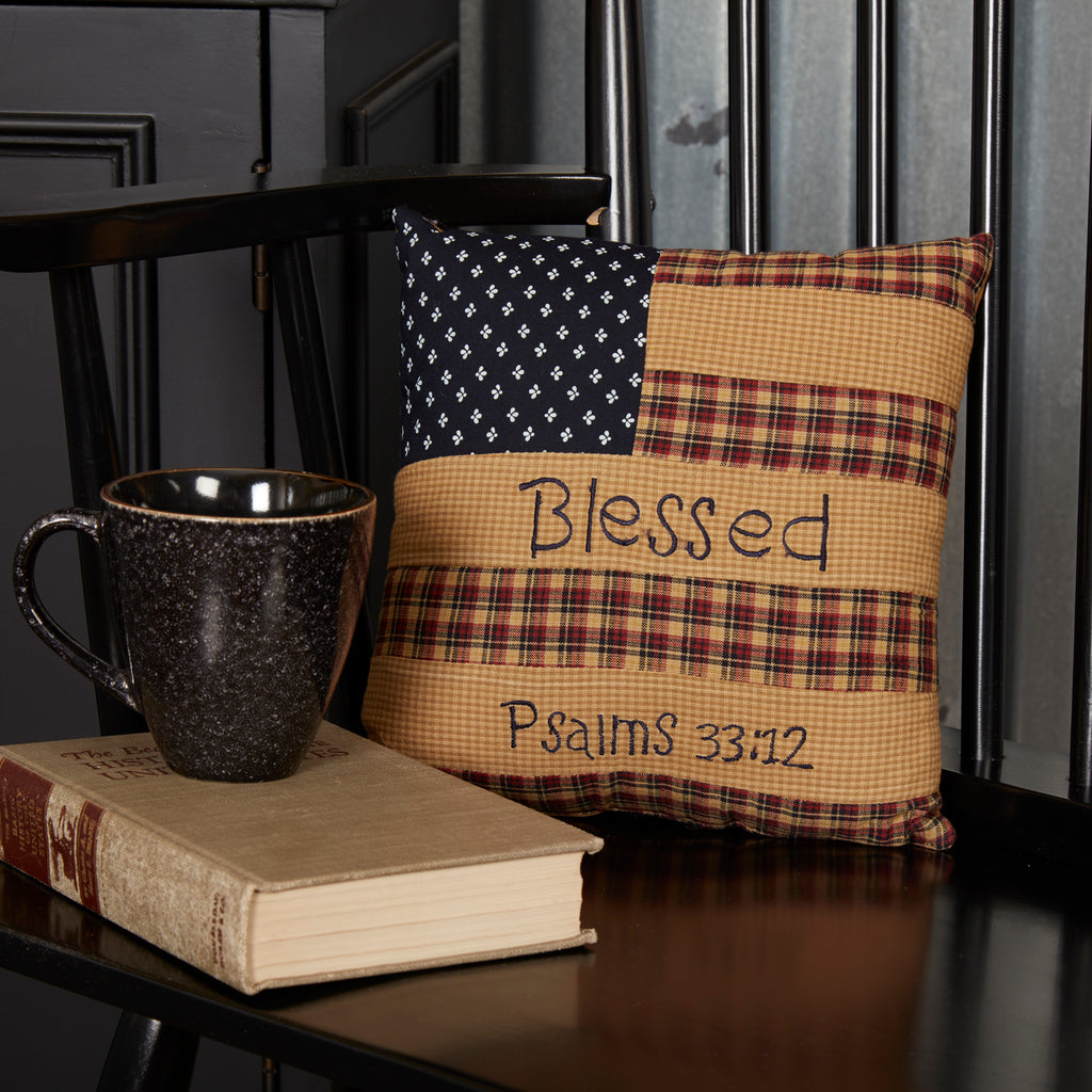 Primitive style 10x10 square accent pillow featuring a tea-stained American flag design, the script word "Blessed," and "Psalms 33:12" printed on a rustic tan background next to a cozy coffee mug setup.