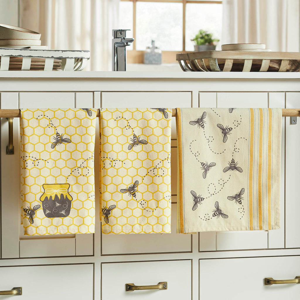 Three kitchen towels with bee and honeycomb patterns hanging on a rack in a kitchen.