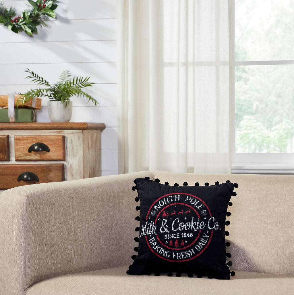 Black Christmas throw pillow with white circular North Pole Milk and Cookie Co logo resting on a light-colored sofa.