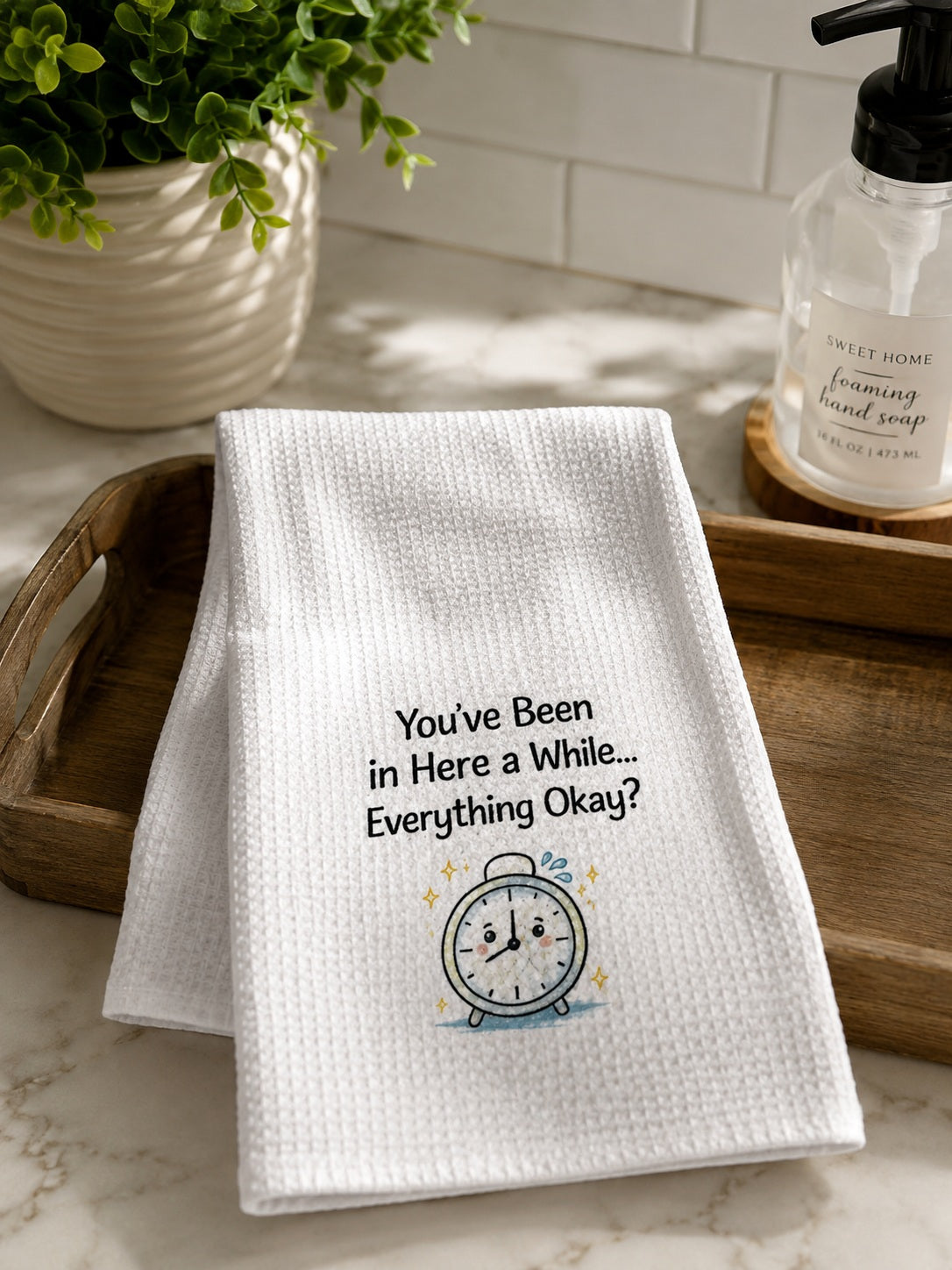 Folded white hand towel displayed in a bathroom setting featuring printed black text reading 'You've Been in Here a While... Everything Okay?' next to a small alarm clock graphic.