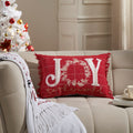 Red holiday accent pillow featured on a sofa with embroidered white "JOY" lettering and a Christmas tree in the background.