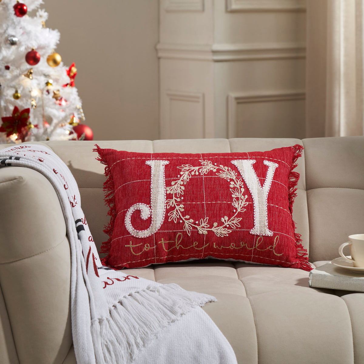 Red holiday accent pillow featured on a sofa with embroidered white "JOY" lettering and a Christmas tree in the background.