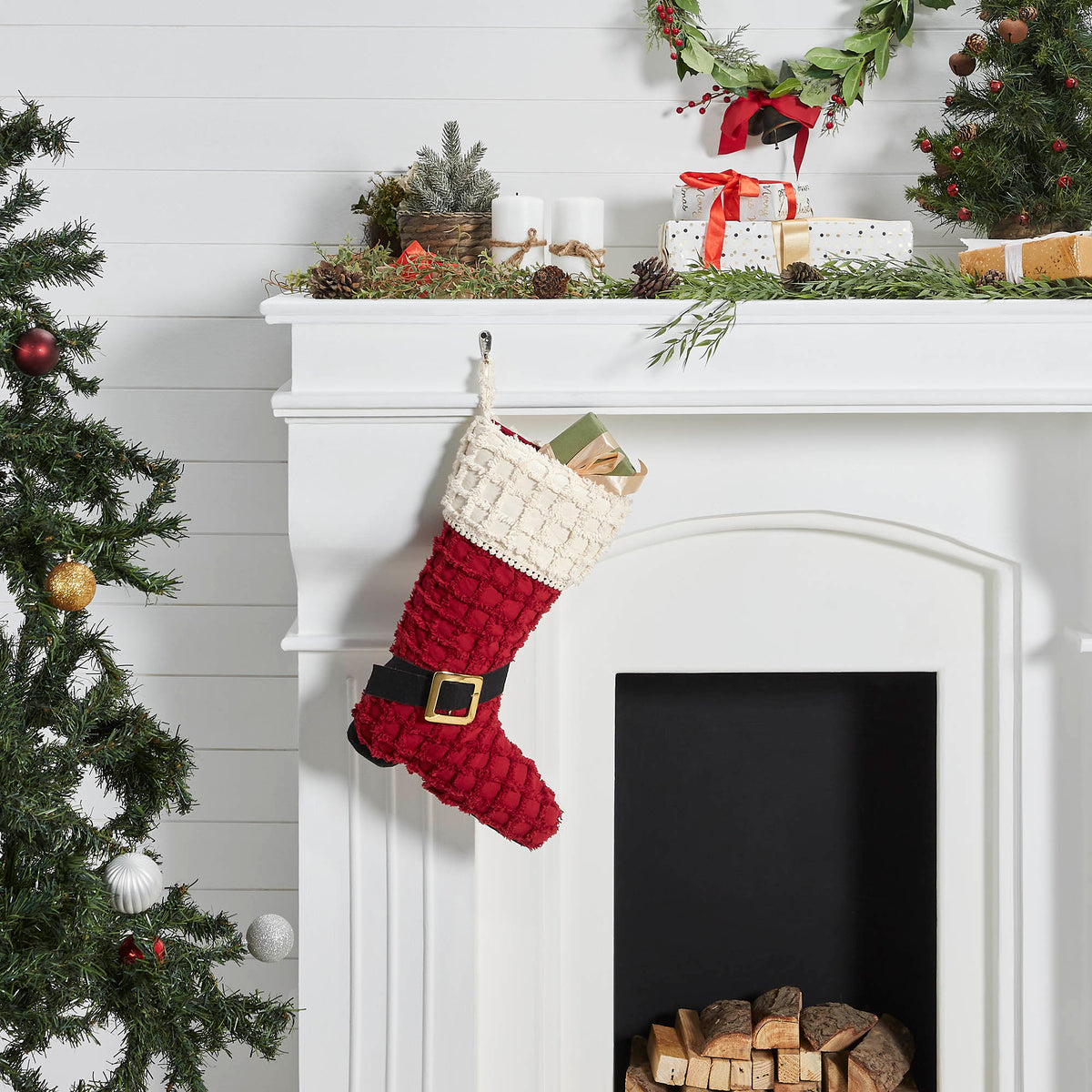 Red and cream knitted Santa boot Christmas stocking hanging on a white fireplace mantel next to a decorated Christmas tree.