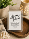 Clear clam-shell package of Mahogany Teakwood luxury soy wax melt on a wooden tray next to a neutral ceramic wax warmer and small potted plant.