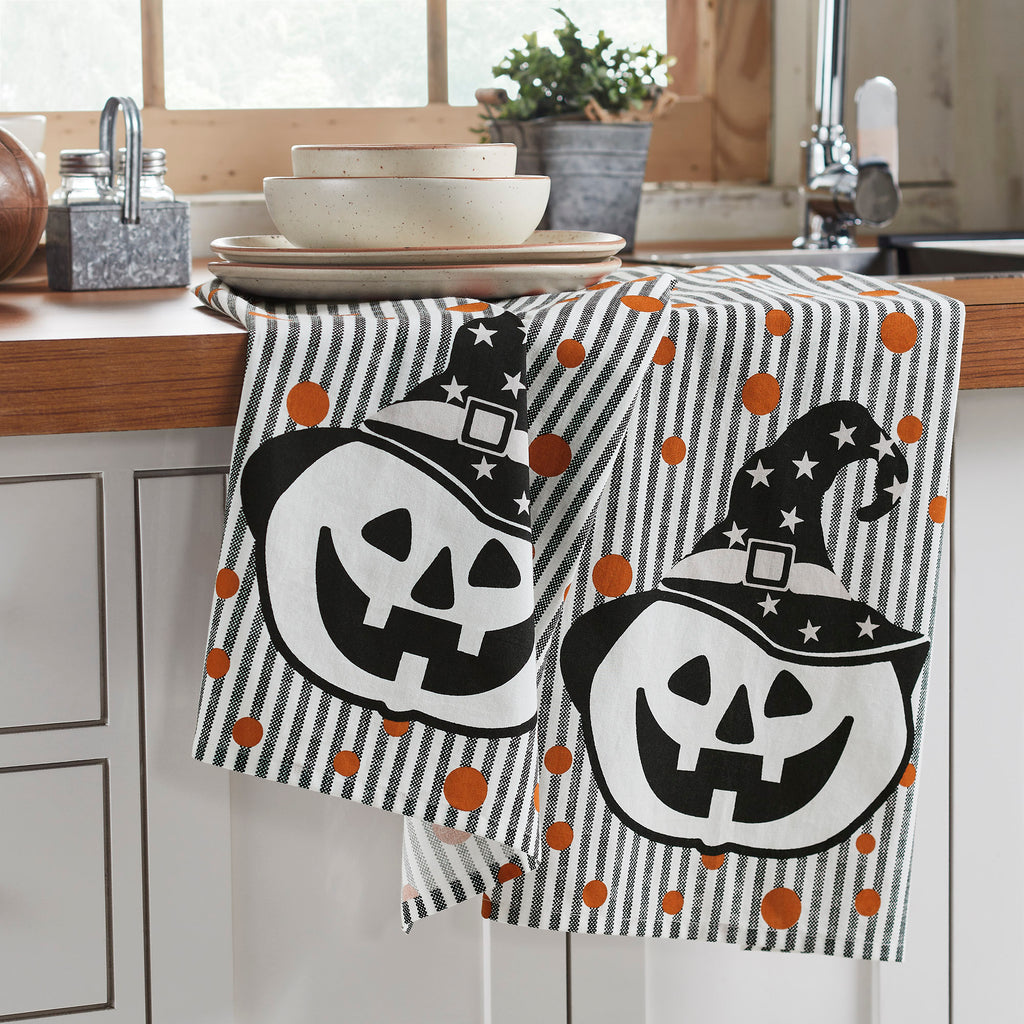 Set of two Halloween kitchen towels with jack-o'-lanterns wearing witch hats displayed on a kitchen counter.