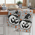 Set of two Halloween kitchen towels with jack-o'-lanterns wearing witch hats displayed on a kitchen counter.