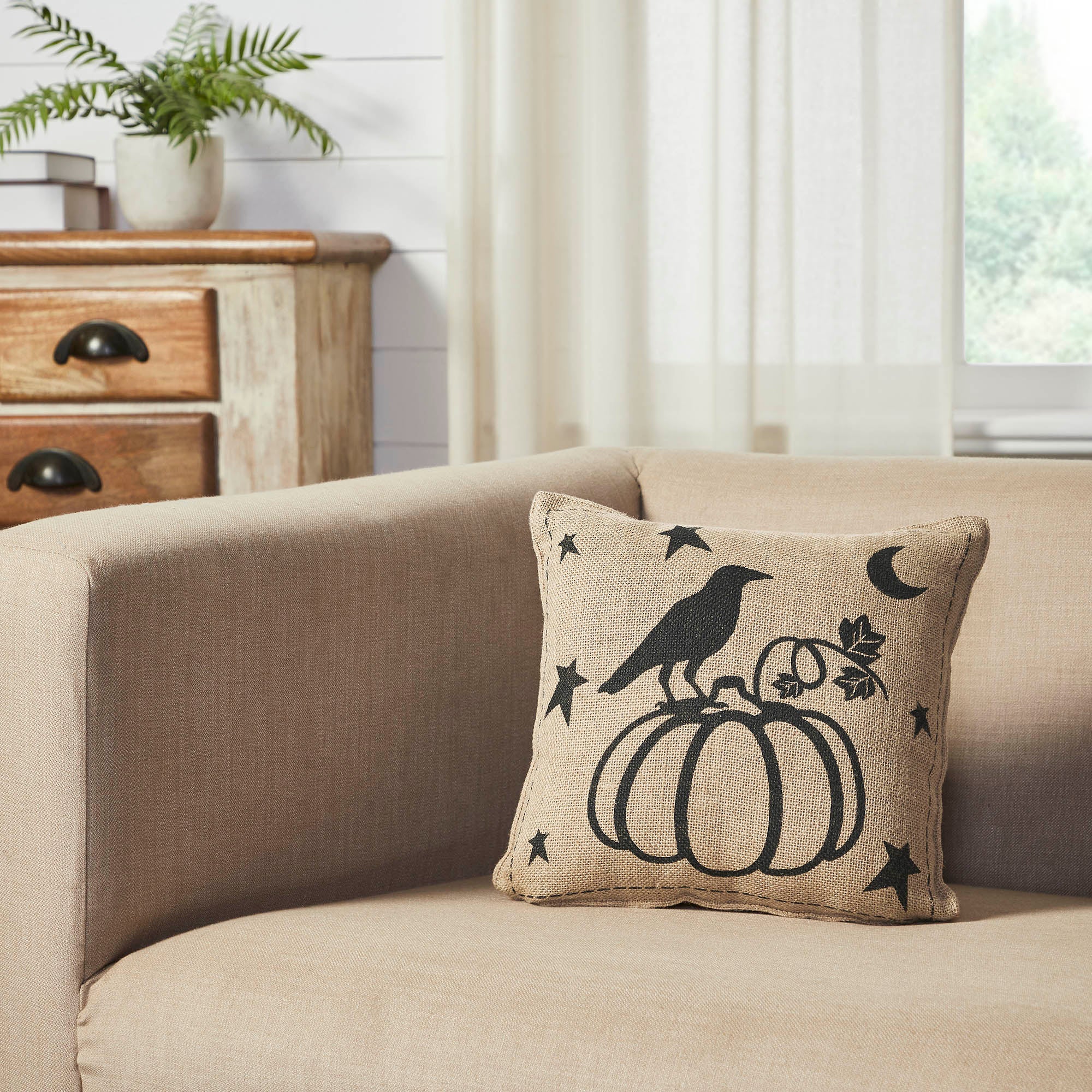 Beige raven and pumpkin throw pillow displayed on a light neutral sofa next to a potted plant.
