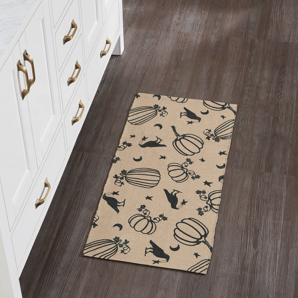 Long rectangular beige area rug with black ravens and pumpkins placed on dark wood floor next to white kitchen cabinets.