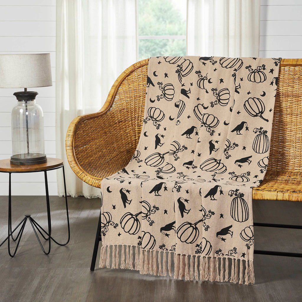 Midnight Harvest woven throw blanket with black ravens, pumpkins, stars and moons draped over a wicker bench in a bright living room.