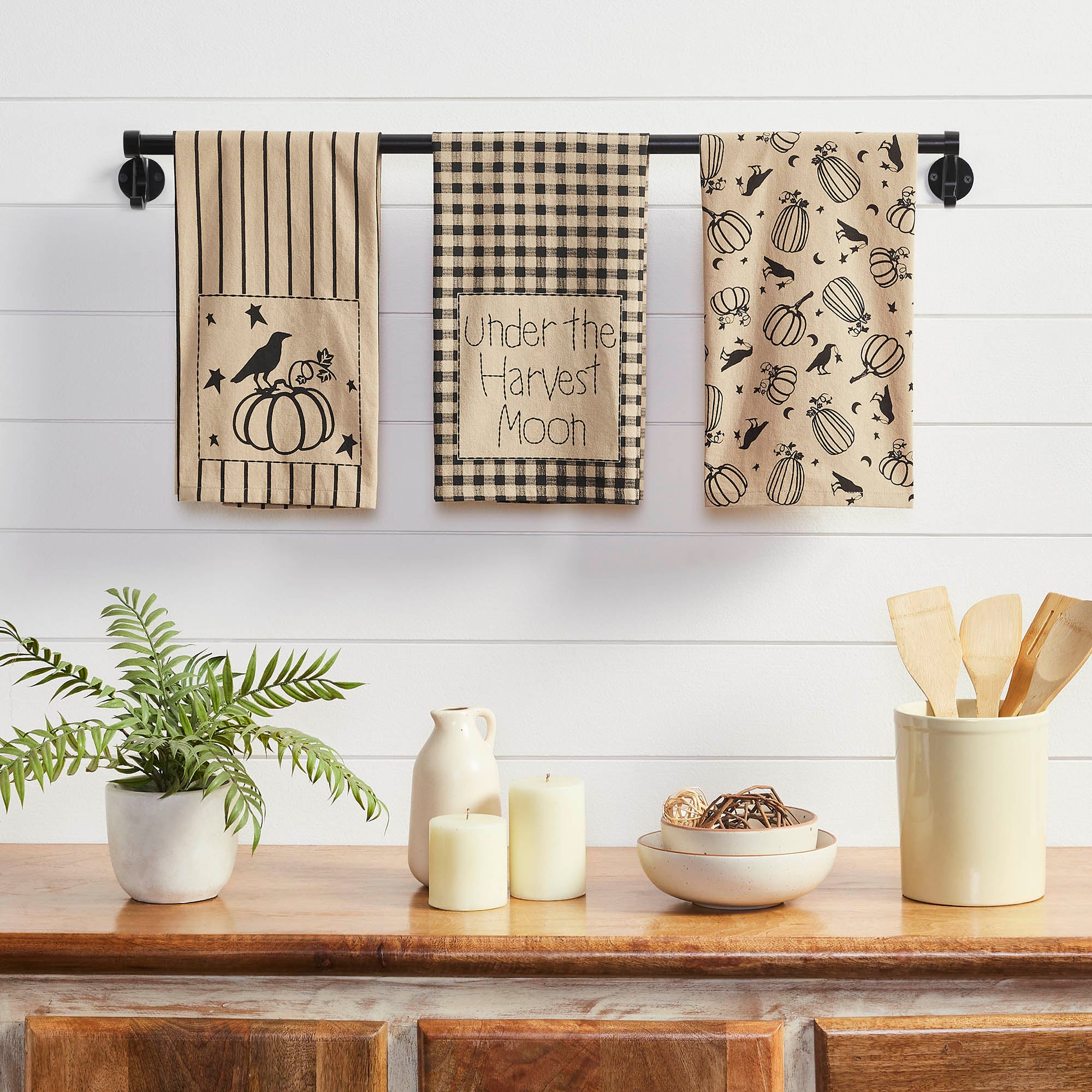 Set of three fall kitchen towels featuring pumpkins, ravens, gingham, grain sack stripes and "Under the Harvest Moon" text displayed on a black wall-mounted towel bar.