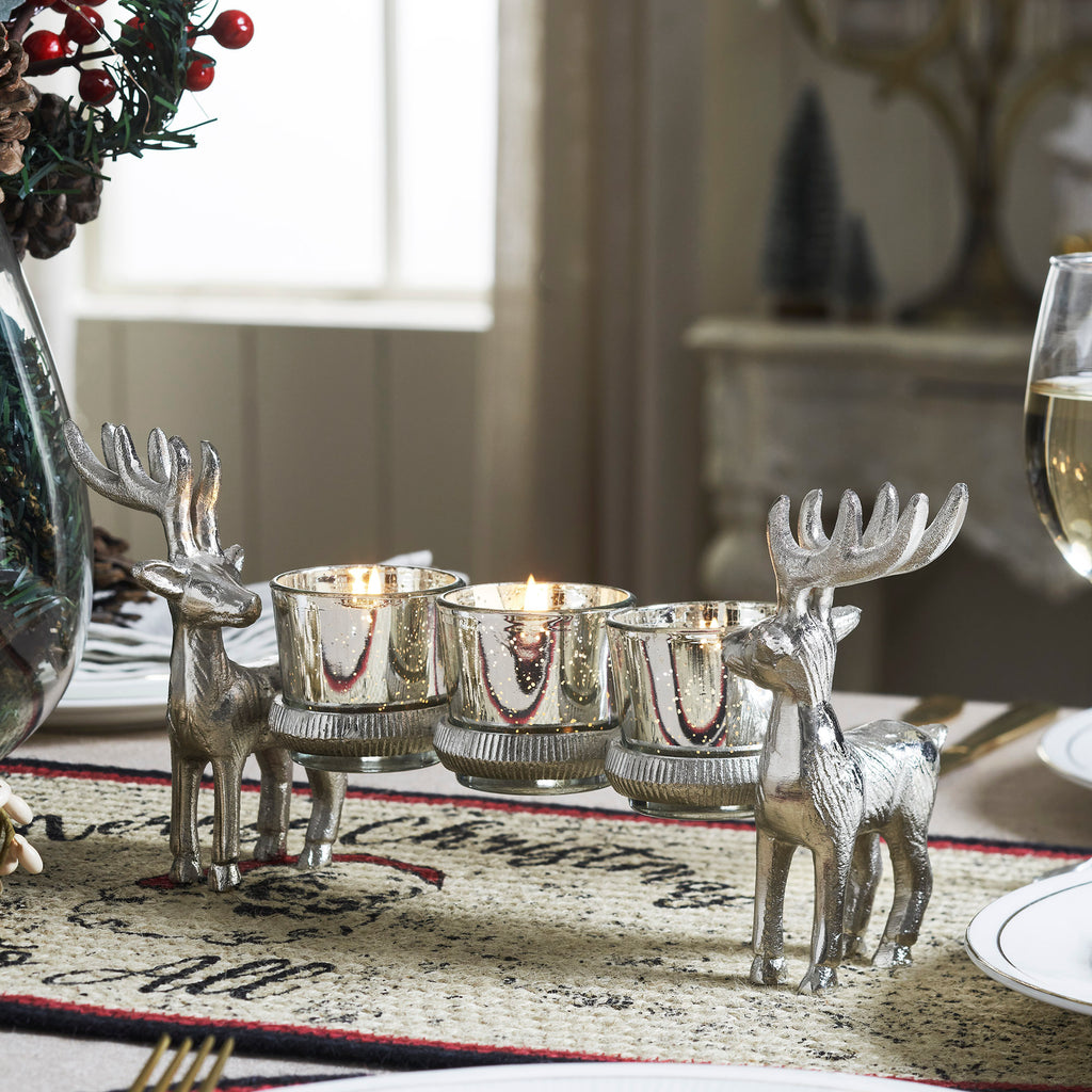 Silver reindeer tealight candle holder with lit candles displayed on a holiday dining table centerpiece.

Image 2 (Front View): Close-up isolated view of the rustic silver metal reindeer candle holder showing four clear glass tealight cups.