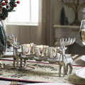 Silver reindeer tealight candle holder with lit candles displayed on a holiday dining table centerpiece.

Image 2 (Front View): Close-up isolated view of the rustic silver metal reindeer candle holder showing four clear glass tealight cups.