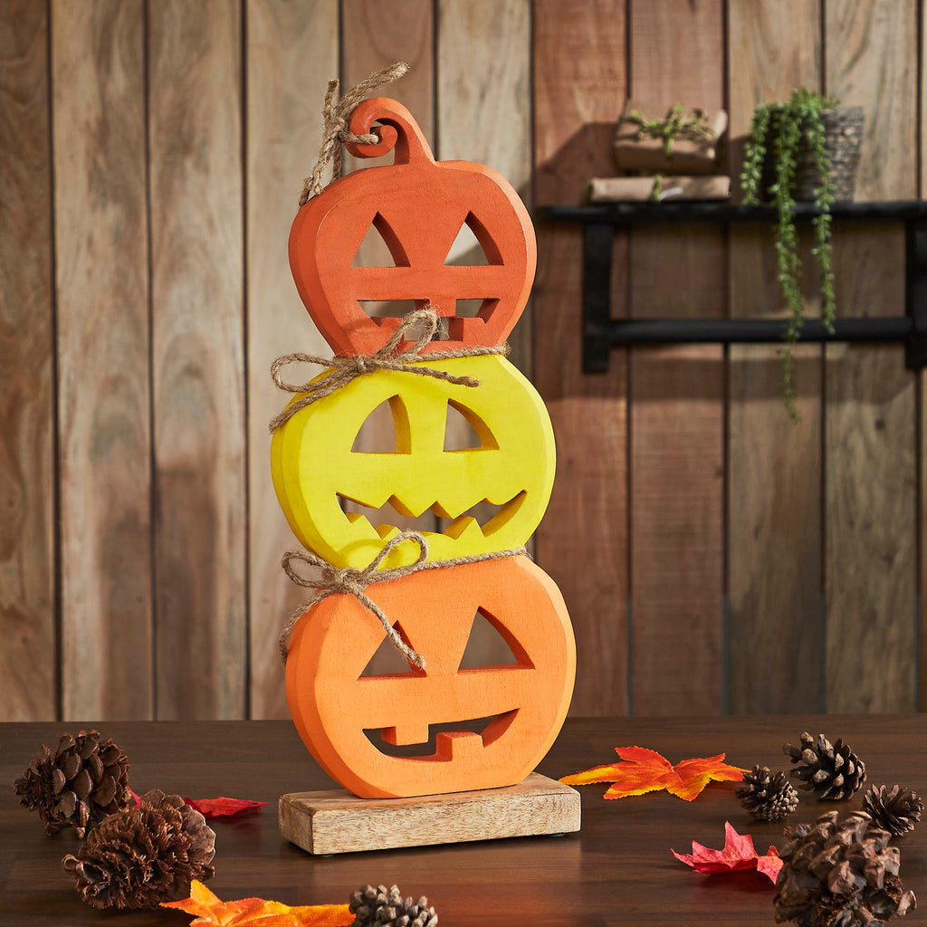 Stacked wooden jack-o'-lantern sign featuring three carved pumpkin faces displayed on a rustic wooden table with fall foliage.