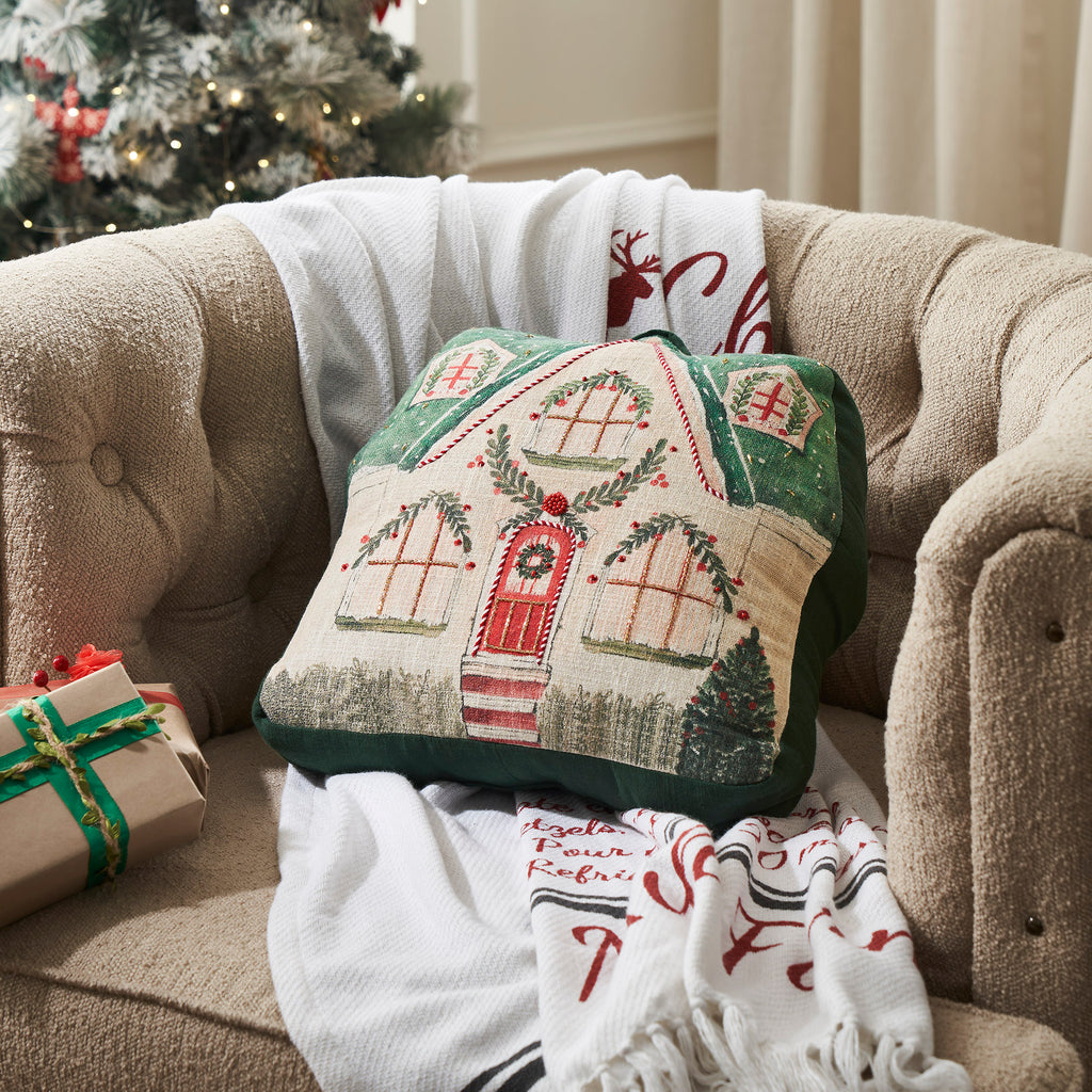 Embroidered Christmas gingerbread house accent pillow styled on an armchair alongside a holiday throw blanket.